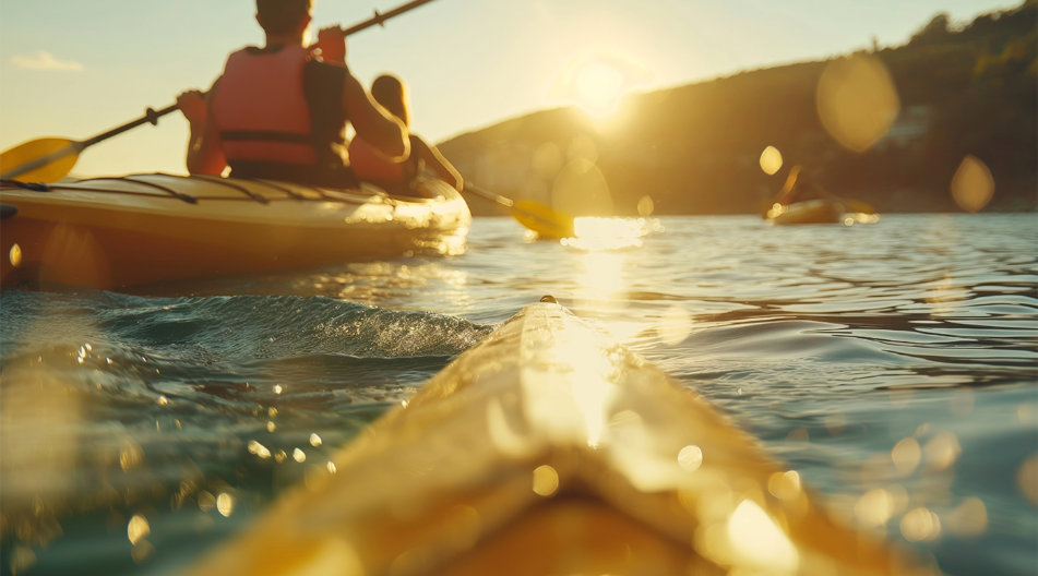 Menschen paddeln in Kajaks bei Sonnenuntergang auf einem ruhigen Gewässer – goldene Lichtreflexe tanzen auf dem Wasser.