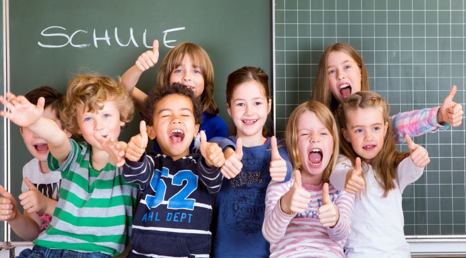 Eine fröhliche Gruppe von Grundschulkindern sitzt vor einer Tafel mit der Aufschrift „Schule“ und zeigt lachend Daumen nach oben.
