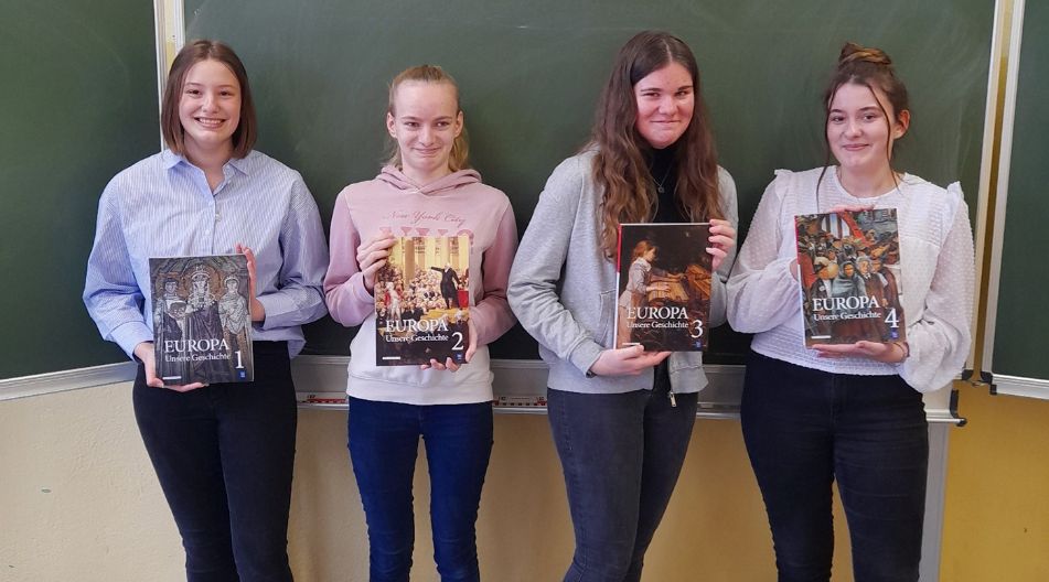 Vier Schülerinnen stehen vor einer Schultafel und halten jeweils einen Band der Geschichtsbuchreihe „Europa – Unsere Geschichte“ (Bände 1 bis 4) in die Kamera. Sie lächeln und präsentieren die Bücher im Klassenraum.
